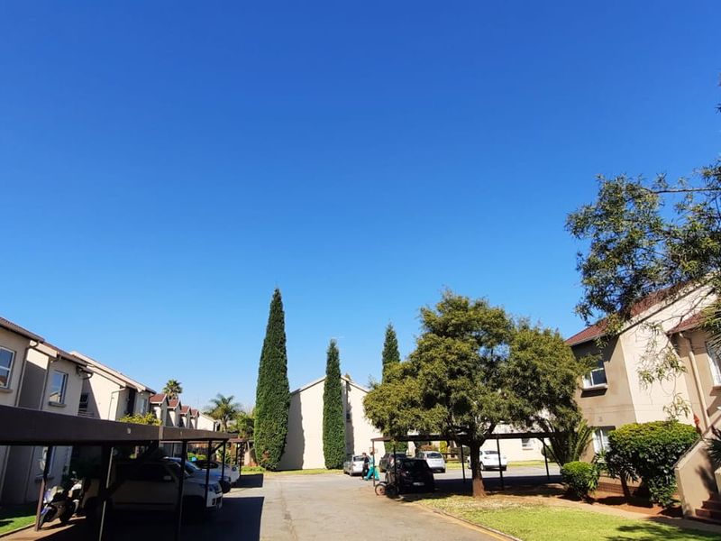 A view of the townhouse complex exterior featuring paved driveways, covered carports, and established garden landscaping under a clear blue sky.