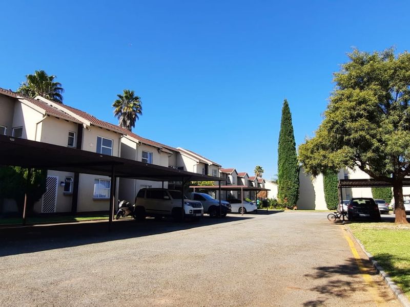 Exterior view of a townhouse complex featuring covered parking bays, paved driveway, and established garden areas under a clear blue sky.