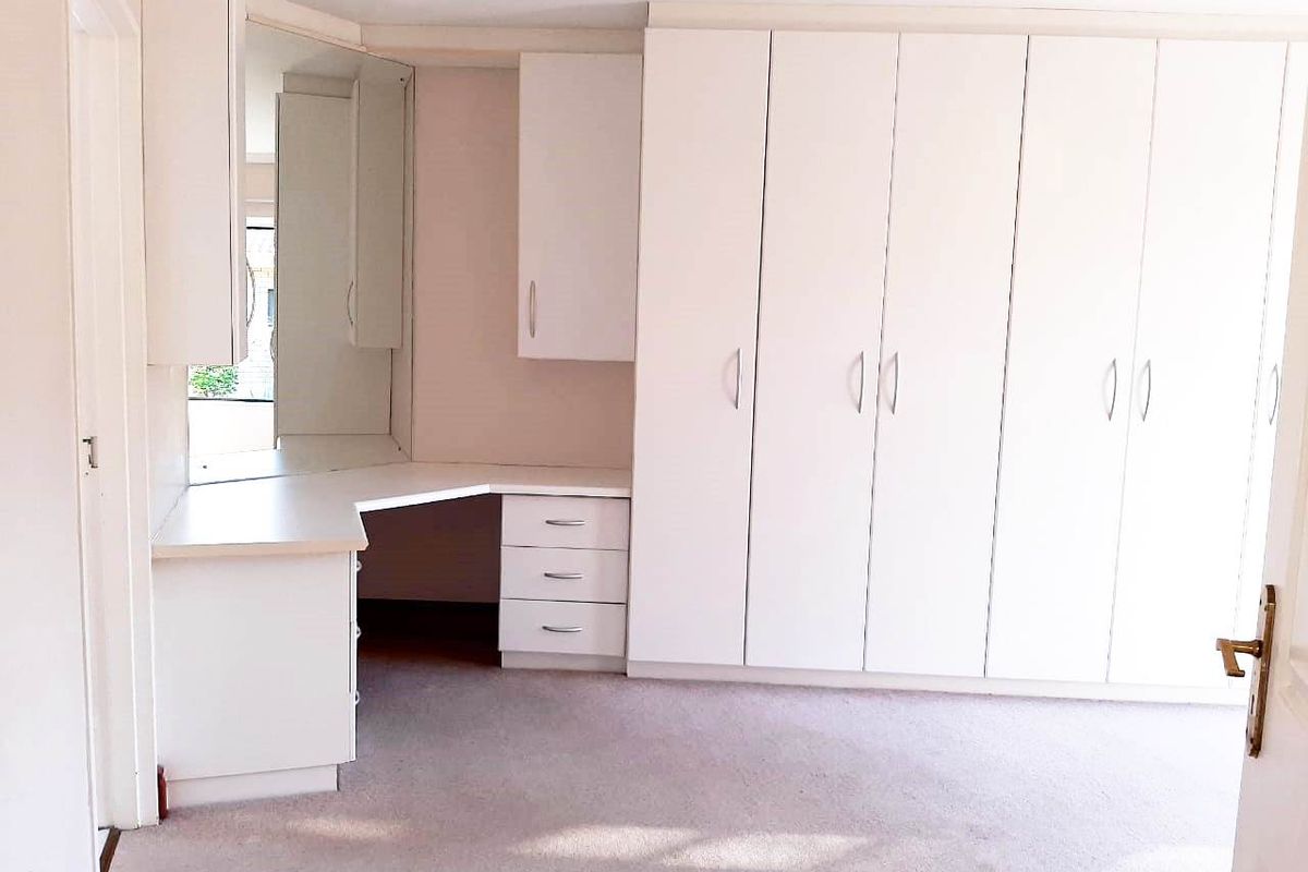 A bright, empty bedroom featuring extensive built-in white cabinetry, a integrated vanity desk with mirror, and neutral carpeted flooring.