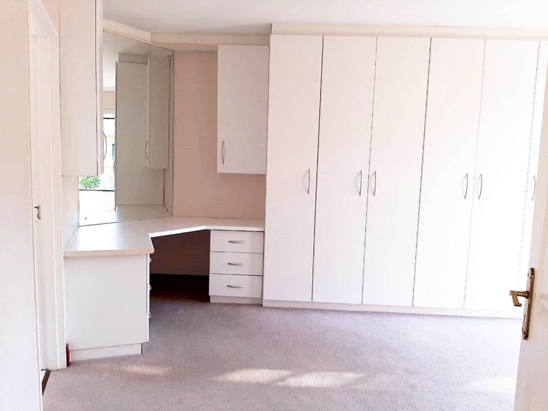 A bright, empty bedroom featuring extensive built-in white cabinetry, a integrated vanity desk with mirror, and neutral carpeted flooring.