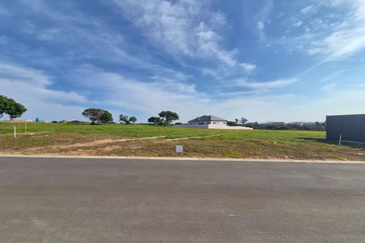 A vacant residential plot of land featuring a level grassed surface, accessible from a paved road in a developed neighborhood.