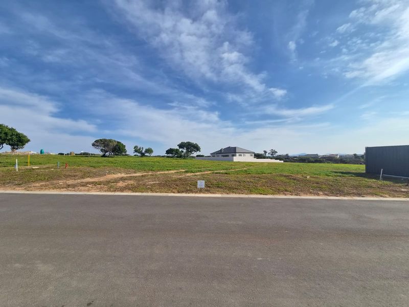 A vacant residential plot of land featuring a level grassed surface, accessible from a paved road in a developed neighborhood.