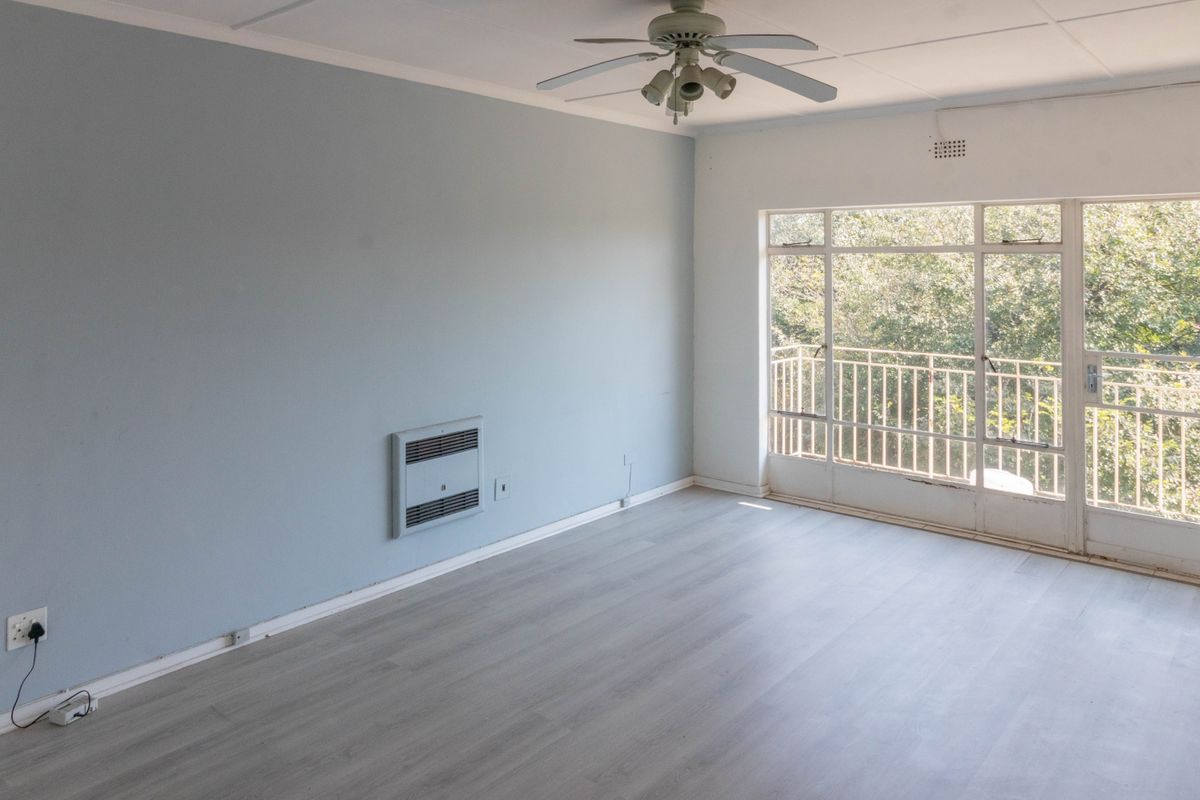 A bright, empty living room featuring light laminate flooring, a ceiling fan, wall-mounted heater, and direct access to a balcony with leafy views.