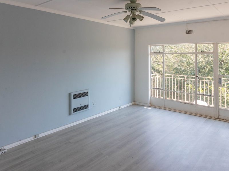 A bright, empty living room featuring light laminate flooring, a ceiling fan, wall-mounted heater, and direct access to a balcony with leafy views.
