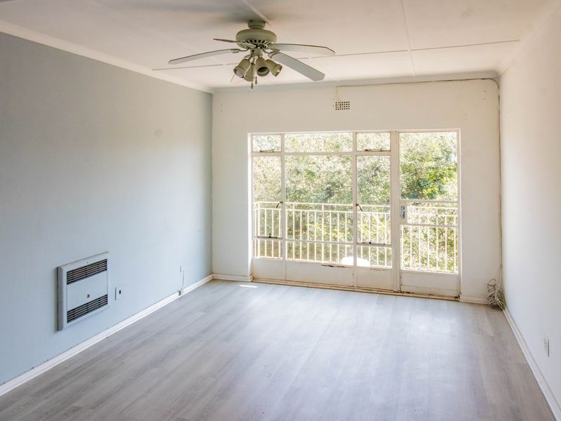 An empty living room featuring light laminate flooring, a ceiling fan, wall-mounted heater, and large glass doors leading to a balcony with leafy views.
