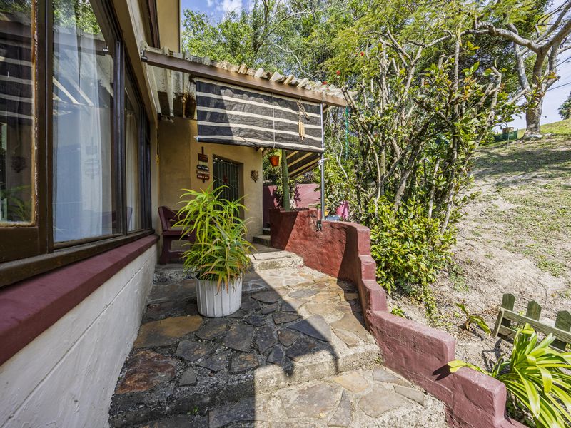 A rustic outdoor patio area featuring stone paving, a covered roof section, and a low retaining wall, set against a sloped garden backdrop.