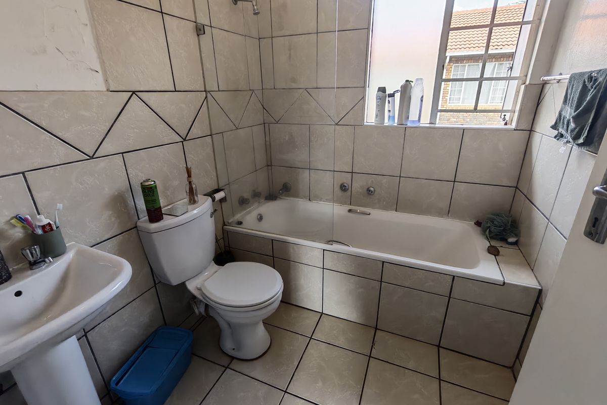 A functional bathroom featuring a pedestal basin, toilet, and a bathtub with a glass shower screen, finished with neutral wall and floor tiles.