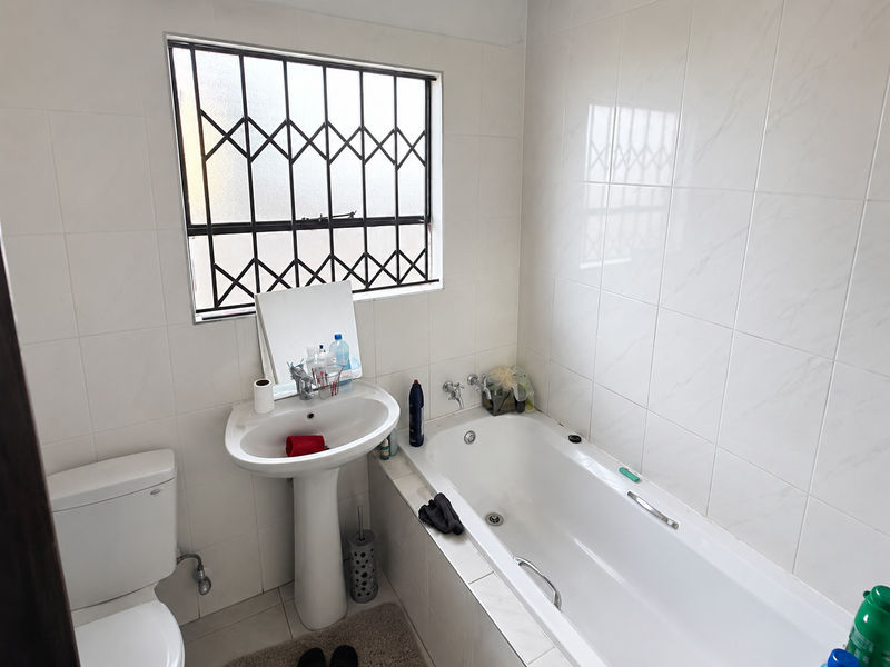 A functional bathroom featuring a pedestal basin, bathtub, and toilet, with tiled walls and a window fitted with a security gate.