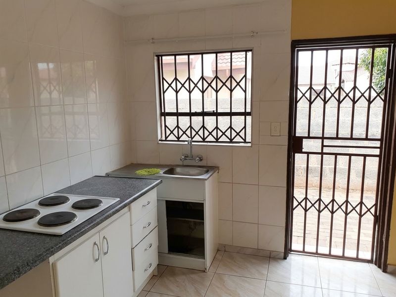 A compact kitchen featuring a built-in electric hob, stainless steel sink, tiled splashbacks, and direct access to the exterior via a security gate.