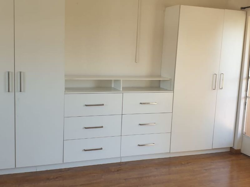A clean, empty bedroom featuring white built-in cupboards with integrated drawer storage and durable laminate flooring, leading to an exterior security door.