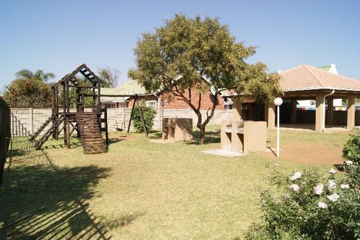 A communal garden area featuring a built-in braai, a covered patio structure, and a children's play area within a fenced residential complex.