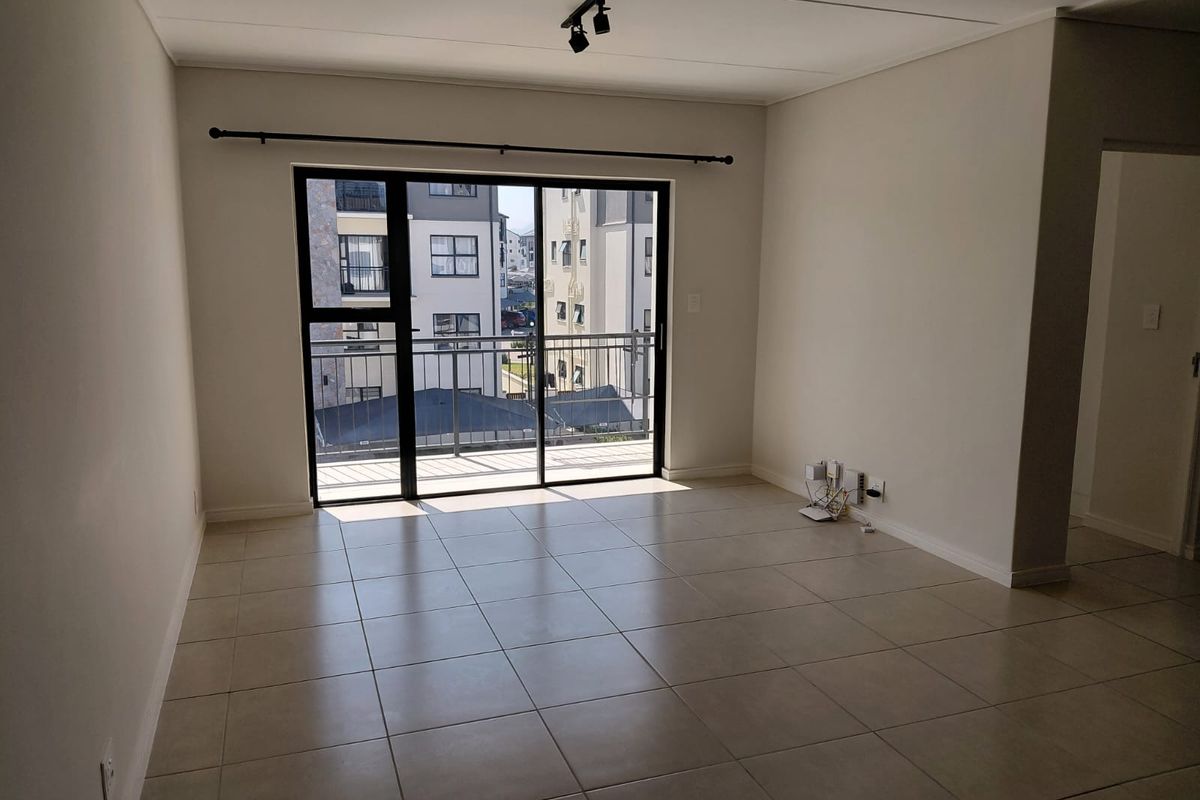 A bright, empty living room featuring neutral tiled flooring, modern track lighting, and a large sliding glass door providing access to a private balcony.