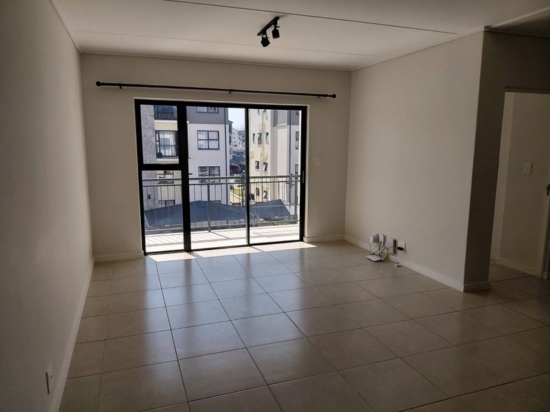 A bright, empty living room featuring neutral tiled flooring, modern track lighting, and a large sliding glass door providing access to a private balcony.