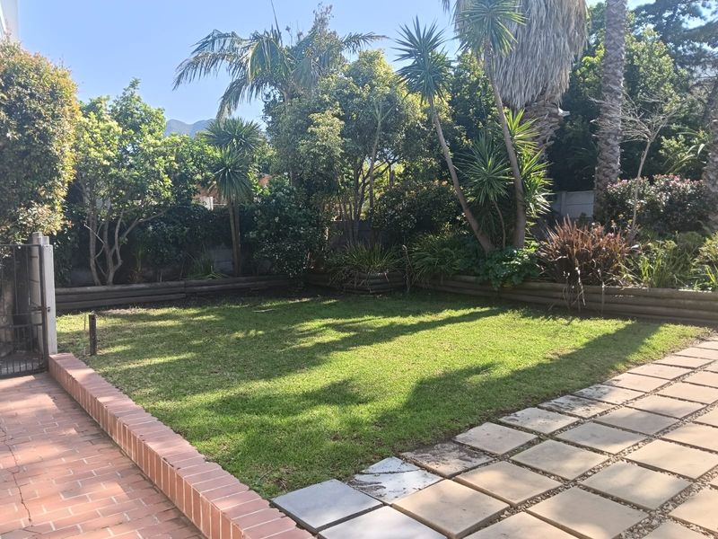 A well-established garden featuring a central lawn, brick retaining walls, and a paved patio area surrounded by mature trees and shrubs.