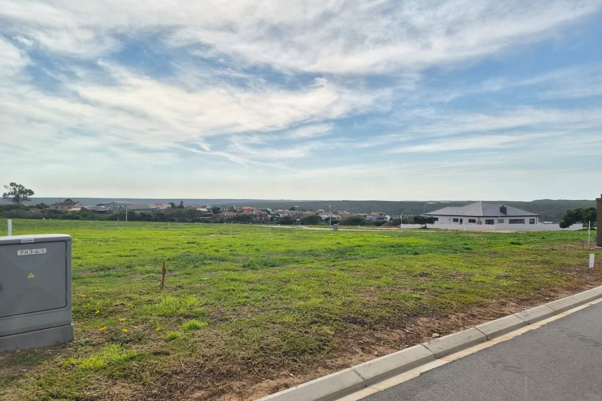A vacant residential plot featuring a level grassy surface, concrete curbing, and an adjacent asphalt road with an electrical distribution box on-site.