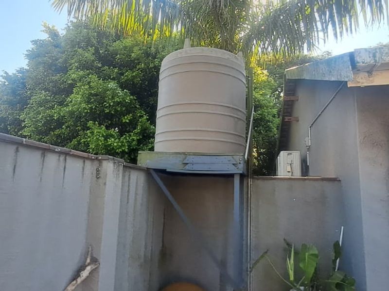 Rear exterior area featuring a water storage tank on a metal stand, boundary wall, and building drainage infrastructure.