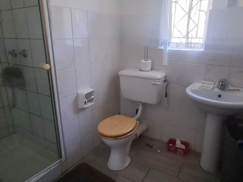A functional bathroom featuring a shower enclosure, pedestal basin, and toilet with tiled wall finishes and security bars on the window.