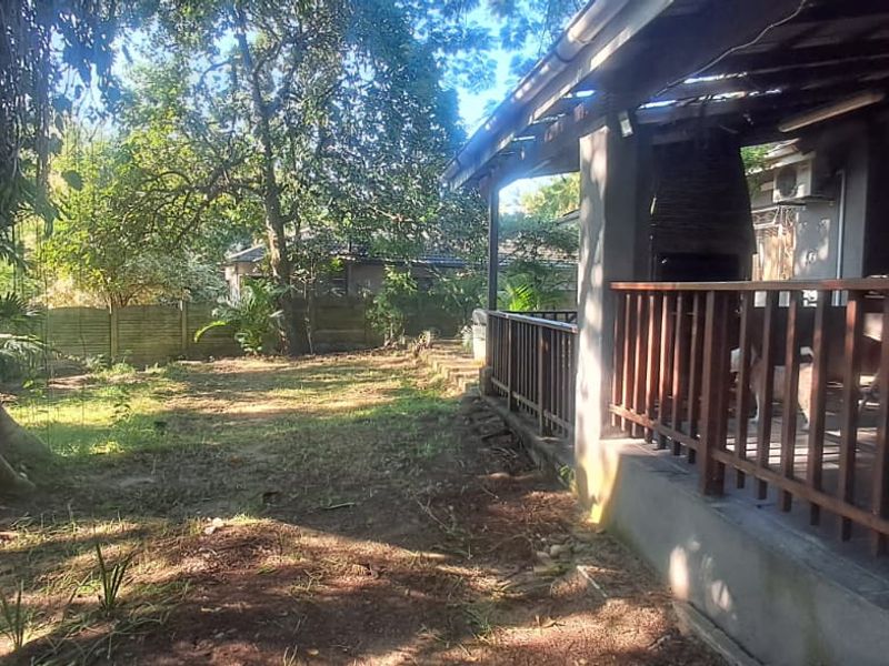 Rear garden area featuring a covered patio with a built-in braai and timber balustrade, overlooking a spacious backyard with mature trees.