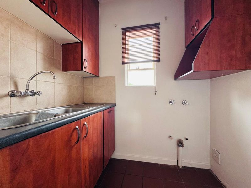 A functional laundry or scullery area featuring built-in wooden cabinetry, a stainless steel sink, tiled backsplash, and plumbing connections for appliances.