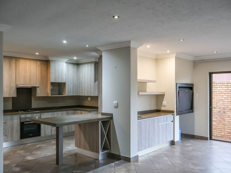Modern open-plan kitchen featuring wood-finish cabinetry, stone countertops, integrated appliances, and a built-in braai area, leading to an outdoor space.