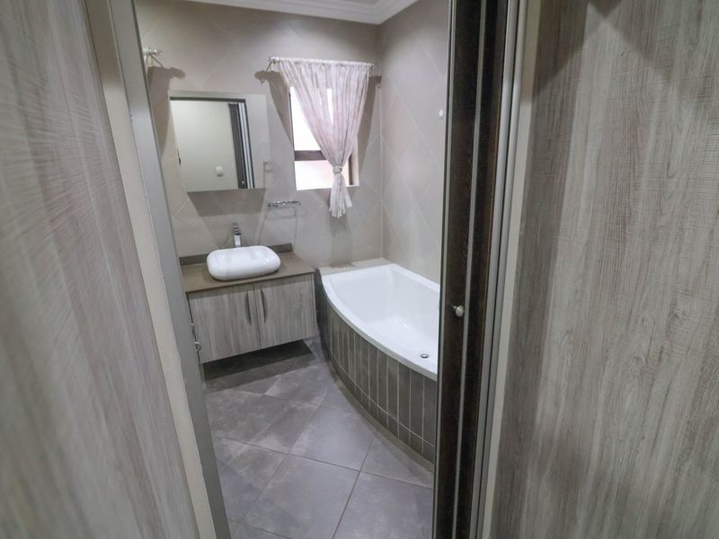 A clean, modern bathroom featuring a built-in vanity with a vessel sink, a bathtub with tiled surround, and neutral wall and floor tiles.