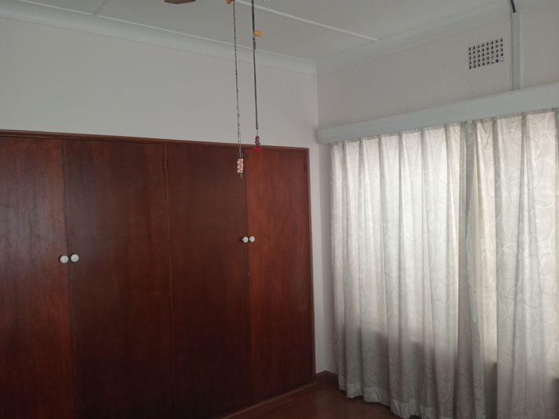 A bedroom featuring a large built-in wooden wardrobe, ceiling fan, and window with curtains.