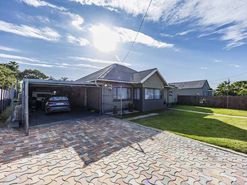 Front view of a residential property featuring a paved driveway, attached carport, and a well-maintained front lawn with perimeter boundary walls.