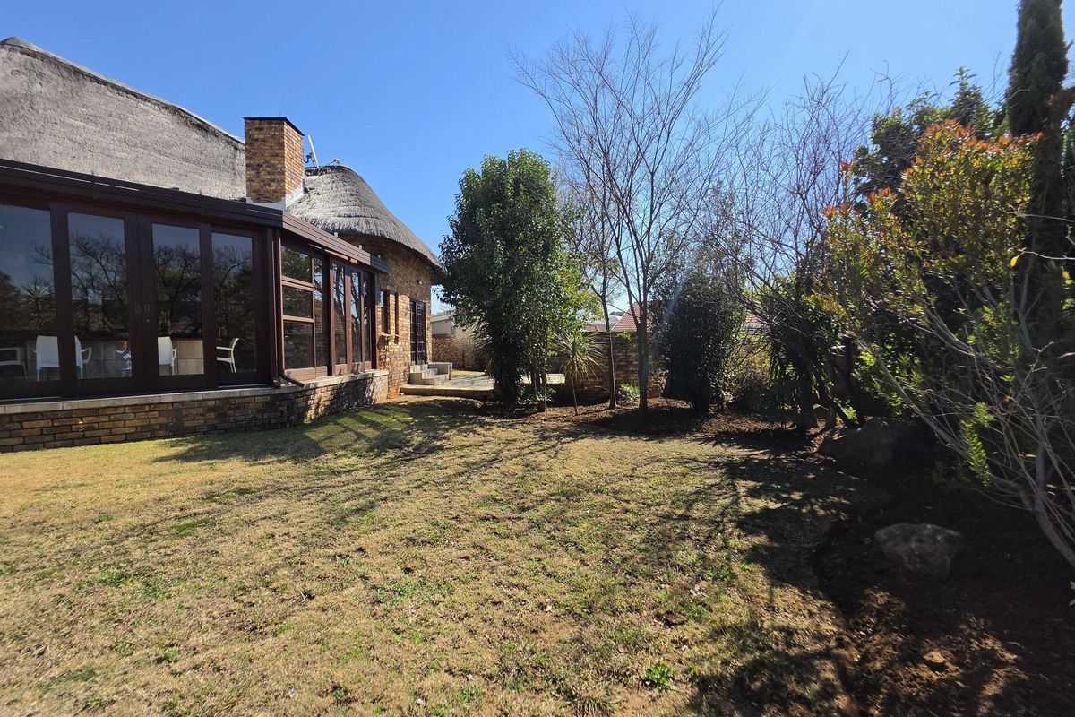 Rear view of a thatch-roof house featuring large glass sliding doors, a brick chimney, and a spacious garden with established trees and boundary walls.