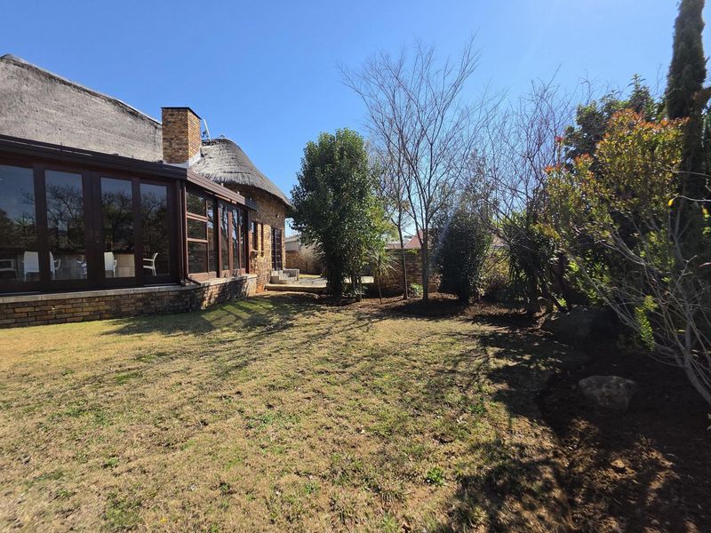 Rear view of a thatch-roof house featuring large glass sliding doors, a brick chimney, and a spacious garden with established trees and boundary walls.