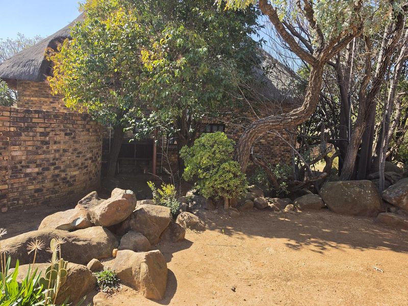 A view of the rear exterior featuring a traditional face brick facade and a thatched roof, surrounded by established trees and natural rock landscaping.