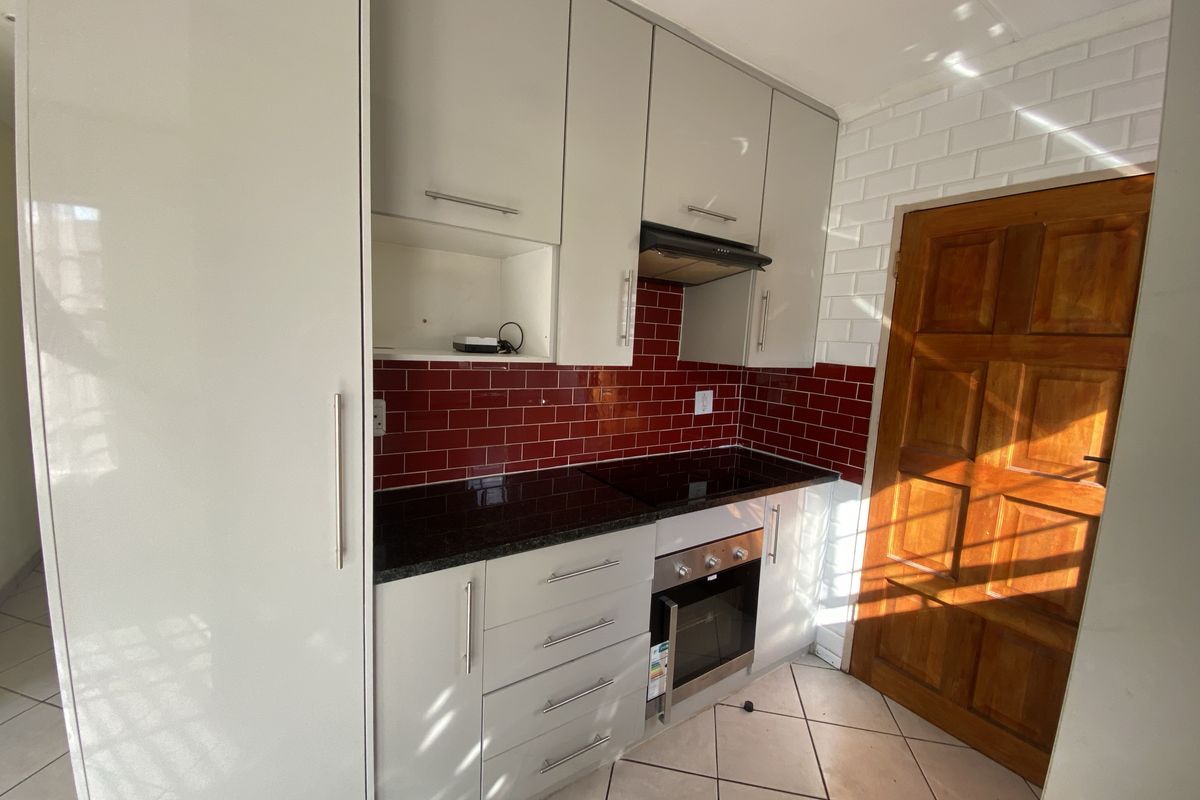 A compact, modern kitchen featuring light grey cabinetry, dark granite countertops, a red subway tile backsplash, and integrated cooking appliances.