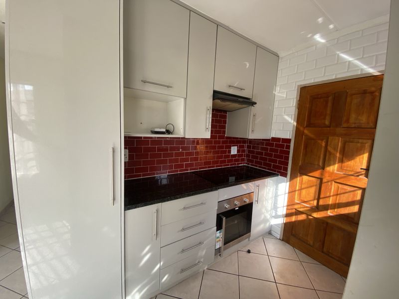 A compact, modern kitchen featuring light grey cabinetry, dark granite countertops, a red subway tile backsplash, and integrated cooking appliances.