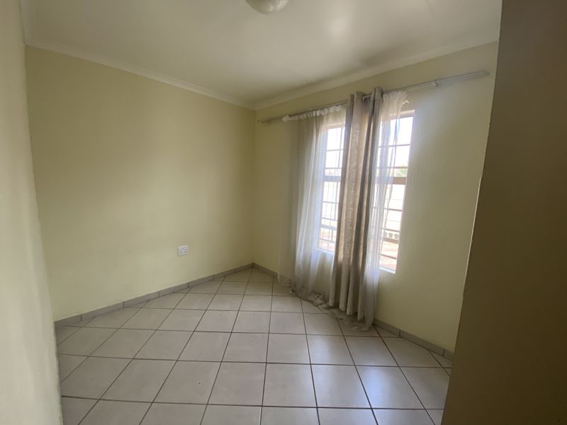 A bright, empty bedroom featuring neutral tiled flooring, coving, and a window fitted with security bars.