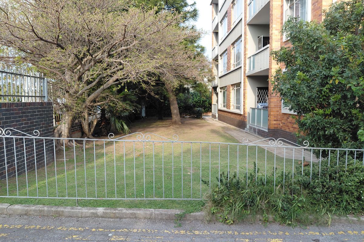 Rear view of the apartment building showing the brick facade, balconies, and a shared garden area with metal perimeter fencing.