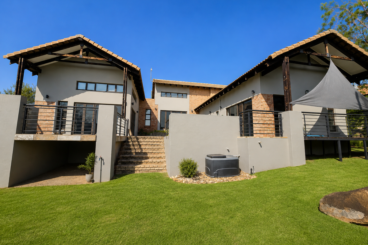 Rear view of a modern house featuring a spacious lawn, raised patios with metal railings, exposed timber roof trusses, and a combination of brick and plaster finishes.