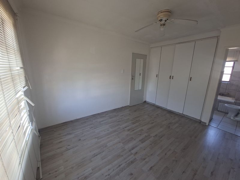A bright, empty bedroom featuring laminate flooring, built-in cupboards, a ceiling fan, and an en-suite bathroom entrance.