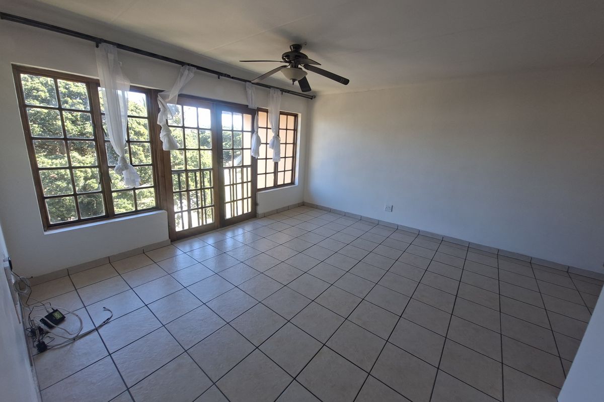 An empty living room featuring tiled flooring, a ceiling fan, and large wooden-framed windows and doors providing natural light.