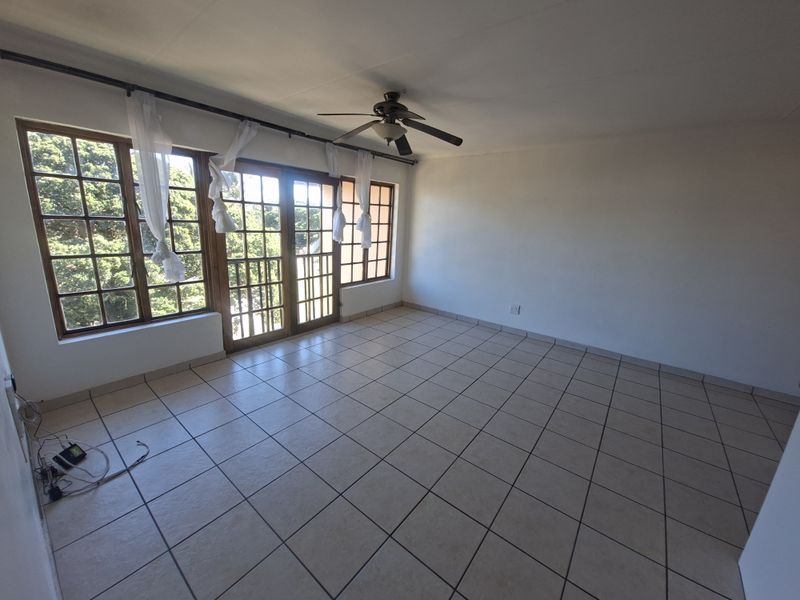 An empty living room featuring tiled flooring, a ceiling fan, and large wooden-framed windows and doors providing natural light.