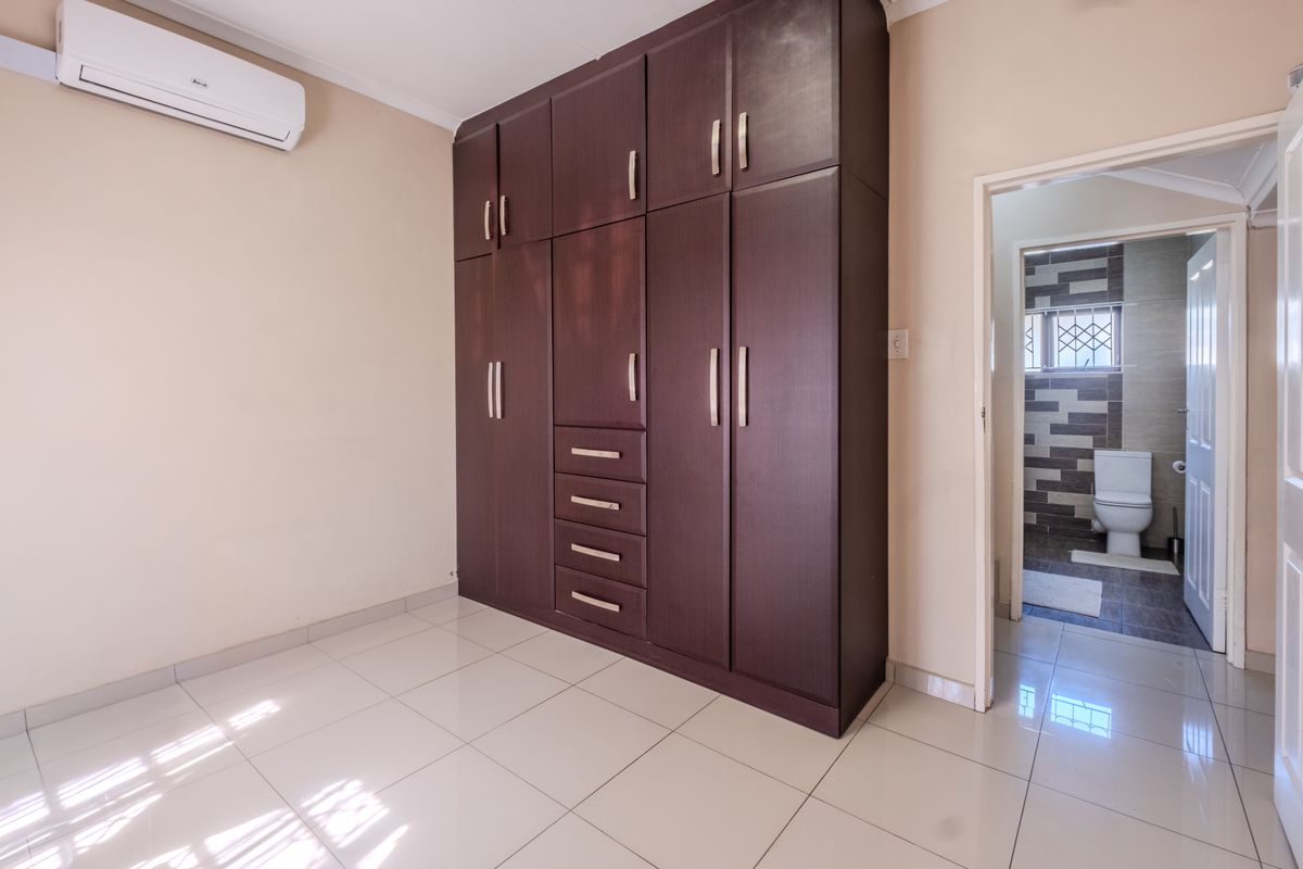 A clean, empty bedroom featuring built-in dark wood cabinetry, tiled flooring, an air conditioning unit, and direct access to an en-suite bathroom.