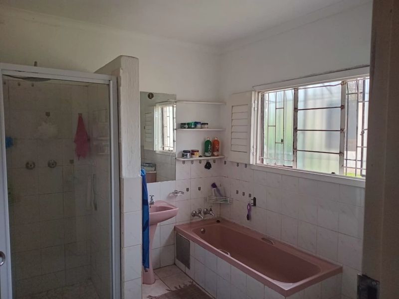A functional bathroom featuring a shower enclosure, a built-in bathtub, and a pedestal basin, with dated finishes and security bars on the window.