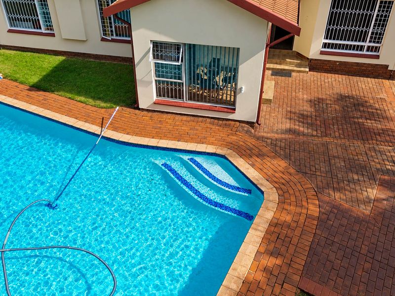An elevated view of a well-maintained swimming pool area featuring brick paving, clear water, and direct access to the home's exterior.