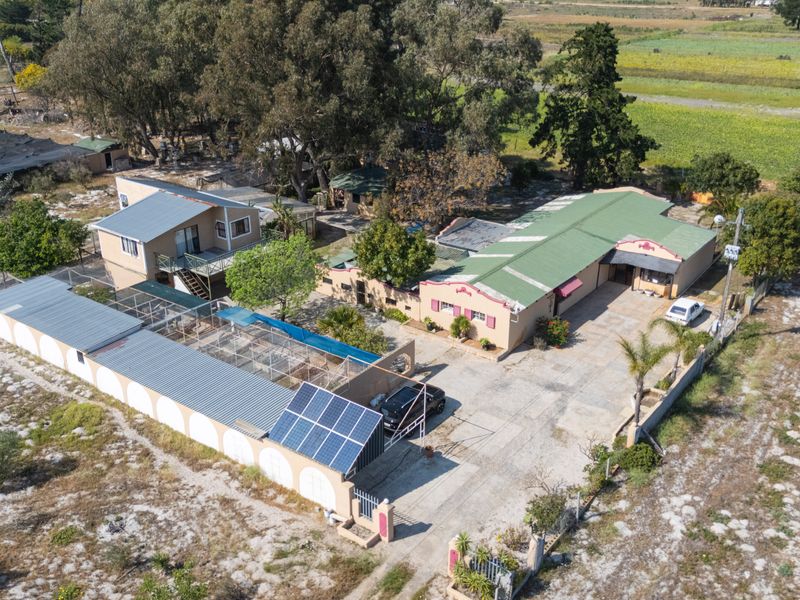 Aerial view of a small holding property featuring multiple structures, solar panel installation, extensive paved driveway, and surrounding rural landscape.