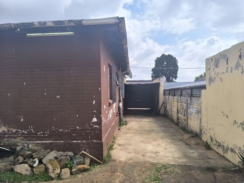Rear exterior view showing a brick house facade, concrete driveway, and boundary wall with visible signs of weathering and paint deterioration.