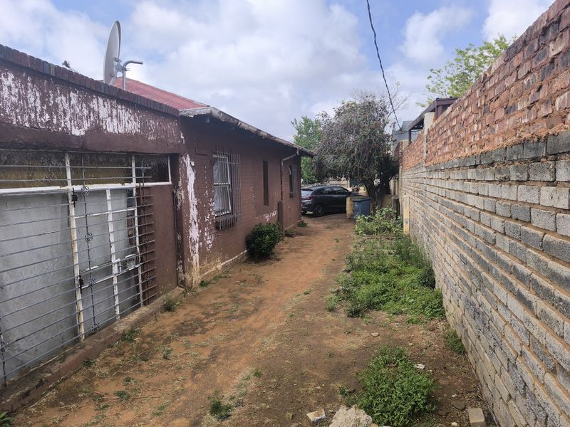 A side exterior view of the property showing a narrow dirt pathway, brick boundary wall, and building facade with security bars.