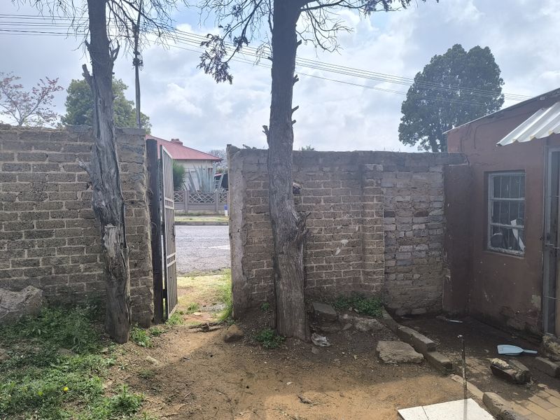 A view of the rear exterior area featuring a brick boundary wall, metal gate, and unpaved ground space.