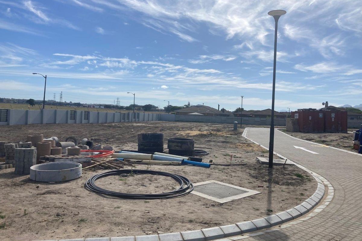 An undeveloped plot of land featuring a paved walkway, perimeter walling, and street lighting, currently used for storage of construction materials.