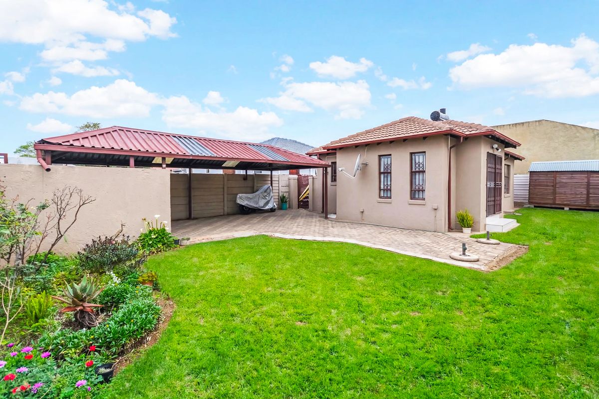 Rear view of the property featuring a paved driveway, a covered carport, a well-maintained lawn, and secure boundary walls.