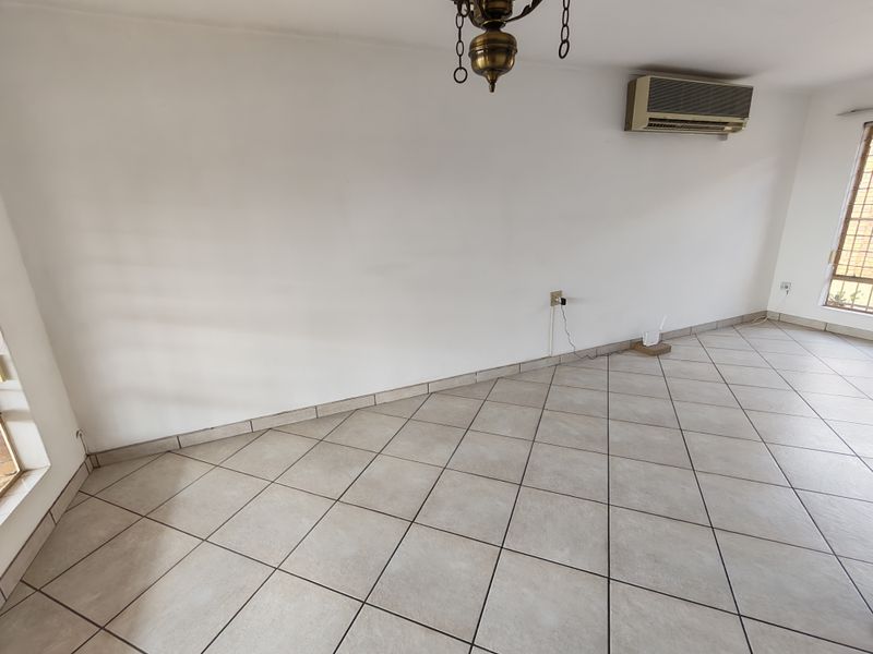 An empty living room featuring neutral ceramic floor tiles, a wall-mounted air conditioning unit, and natural light from large windows.