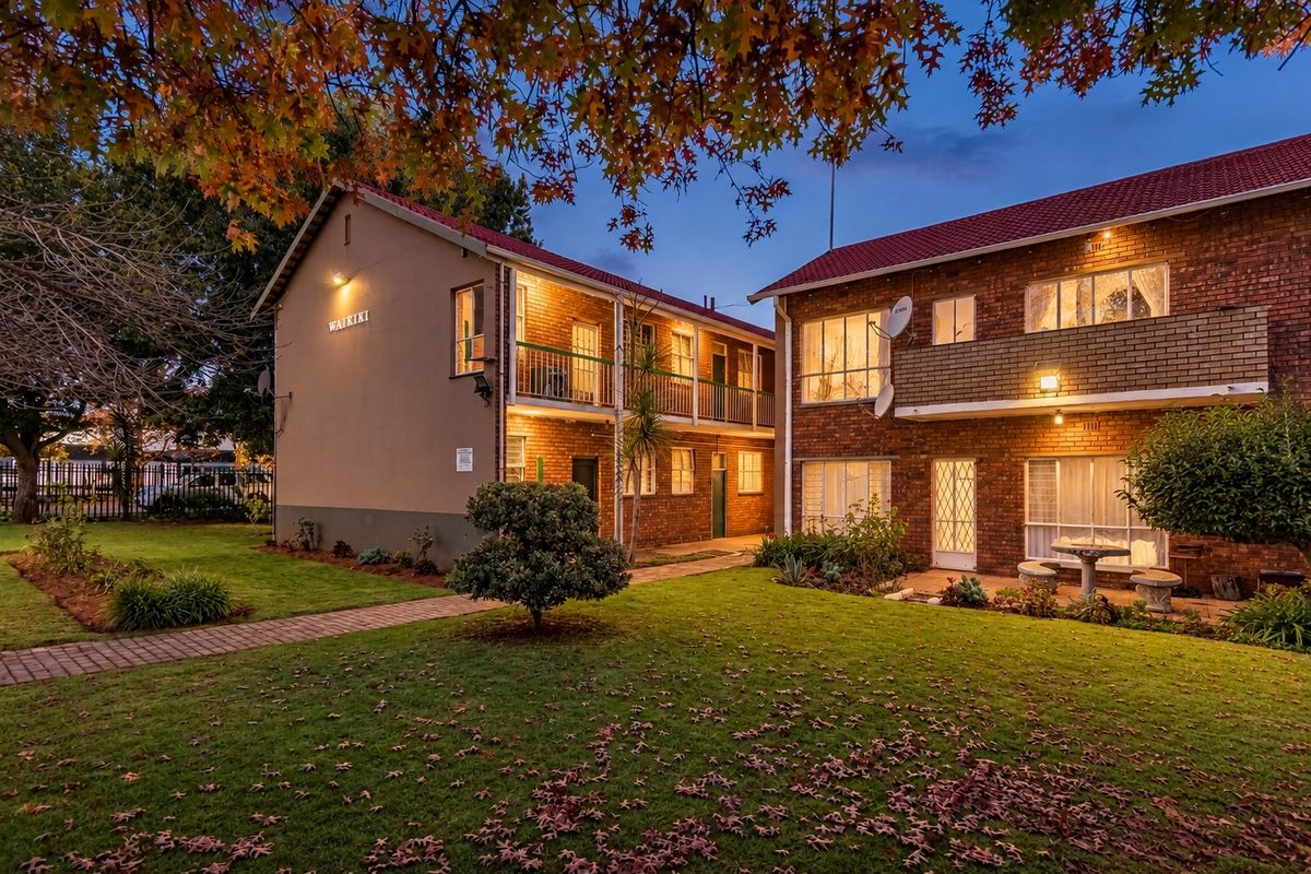 A view of the rear exterior of the Waikiki apartment complex, featuring brick facades, balconies, and a well-maintained communal garden area at dusk.