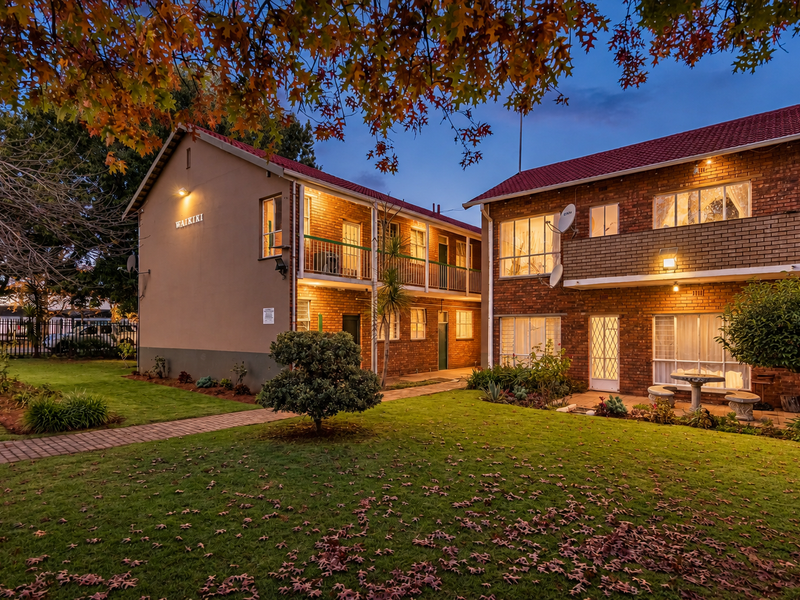 A view of the rear exterior of the Waikiki apartment complex, featuring brick facades, balconies, and a well-maintained communal garden area at dusk.